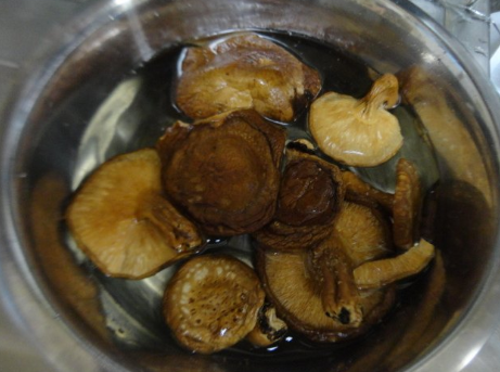 一位厨师在厨房中将泡发好的干香菇放入砂锅中炖煮,蒸汽升腾,周围摆放着葱姜蒜等调料,展现干香菇在实际烹饪中的应用场景 厨房中使用干香菇烹饪的画面