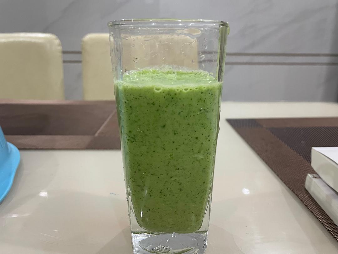 一杯透明玻璃杯中盛有浅绿色的浓稠液体,表面可见细小气泡,杯子旁放有一根吸管和一小把新鲜菠菜叶,背景为木质餐桌和自然光线,营造健康早餐氛围 菠菜酸奶思慕雪成品图