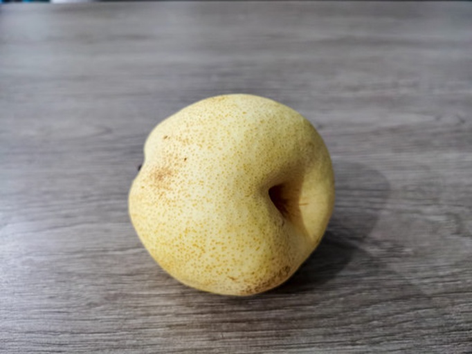 一个硕大的浅黄绿色梨子,果形上宽下窄,顶部宽厚,果柄短粗,表面光滑带有轻微锈斑,放置在木质桌面上,背景为果园环境 大头梨实物图