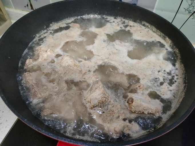 一口黑色铸铁锅正在炉灶上炖煮,锅内可见红褐色的五花肉块与深绿色的豆角混合在一起,汤汁微微冒泡,蒸汽升腾,锅边配有木铲和调料瓶,体现家庭烹饪的真实场景 锅中炖煮中的豆角与五花肉