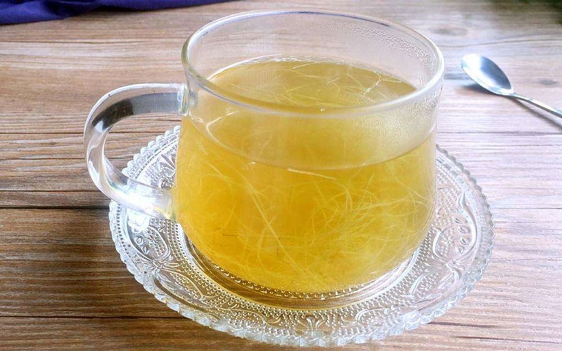 一杯透明玻璃杯中漂浮着3-4片淡黄色参片,水体呈浅金黄色,杯子旁放置一个白色瓷碟,上面有两片干燥参片和一把小银匙,背景为木质桌面和绿色植物 参片泡水饮用实景图