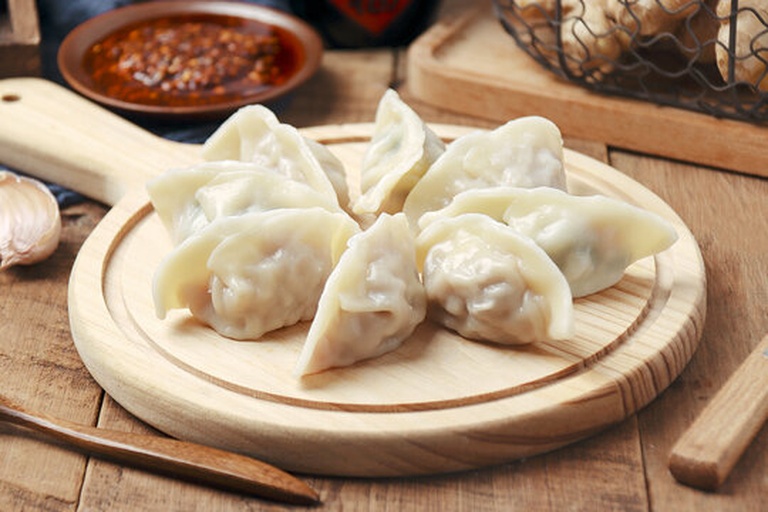一盘刚出锅的彩色圆椒饺子整齐摆放在白色瓷盘中,部分为透明蒸饺皮隐约可见红黄色馅料,旁边配有蘸碟(酱油+香醋+蒜末),背景虚化的餐桌上还有小葱花和辣椒油瓶,整体风格清新诱人 彩色圆椒饺子成品摆盘