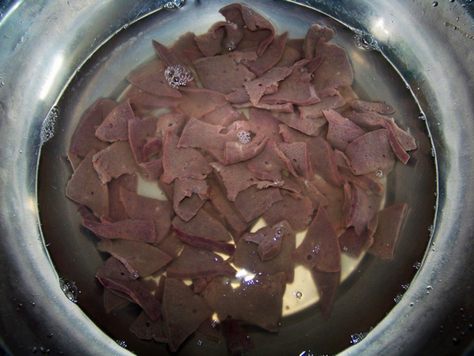 新鲜猪肝被切成均匀薄片,放置在透明玻璃碗中,用清水完全覆盖,水略显淡粉色,表明正在泡出血水。背景为厨房操作台,旁边有砧板和刀具 猪肝切片与清水浸泡过程