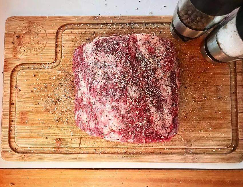 木质餐桌上摆放着不锈钢盐罐和胡椒研磨器,一只手正在旋转研磨器底部,新鲜磨出深褐色胡椒粉洒在牛排表面,旁边配有叉子与餐巾,展现高端料理氛围 厨房中使用胡椒研磨器的场景