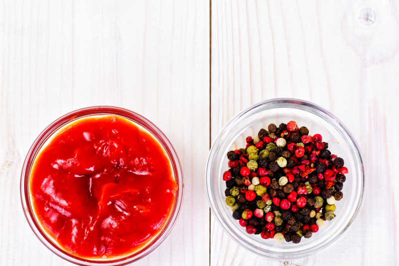 木制托盘上整齐摆放四个玻璃瓶,分别装有黑色胡椒粒、白色胡椒粒、灰色胡椒面和彩色混合胡椒,背景为暖色调厨房环境,展示不同胡椒形态与用途 多种胡椒产品陈列图