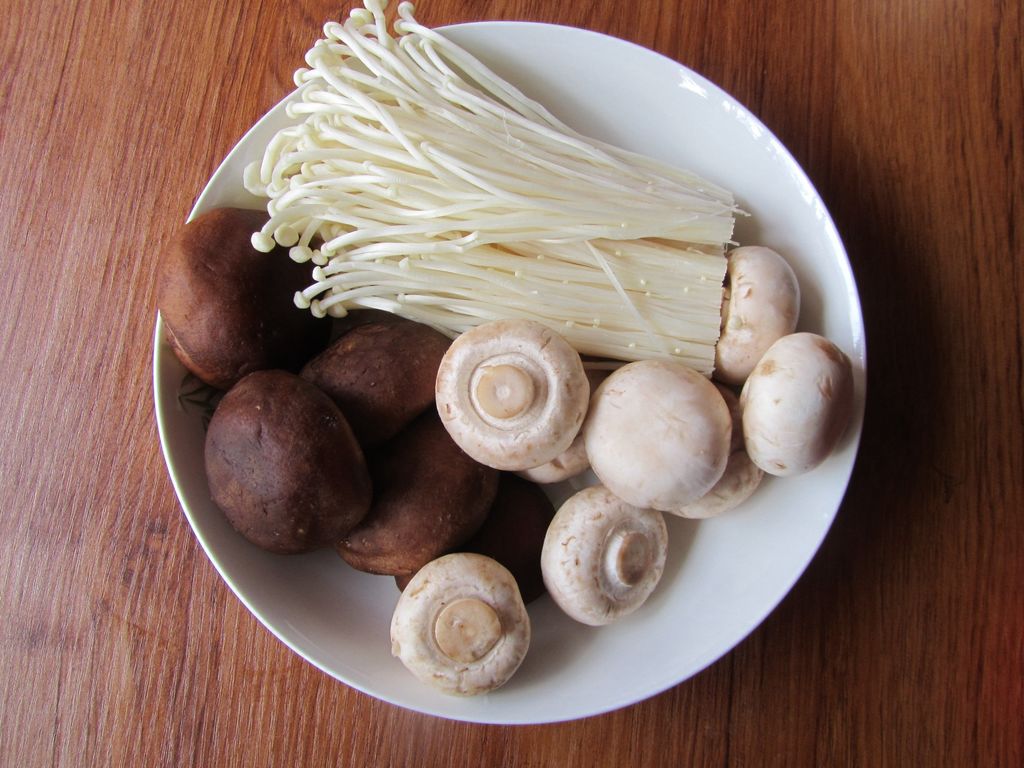 三种菌菇已清洗并处理完毕:左侧为撕成条状的鲜平菇,中间为去根后散开的金针菇,右侧为切成瓣状的白蘑菇,整齐摆放在白色瓷盘中,背景为木质料理台 鲜平菇、金针菇、白蘑菇分别处理后的状态