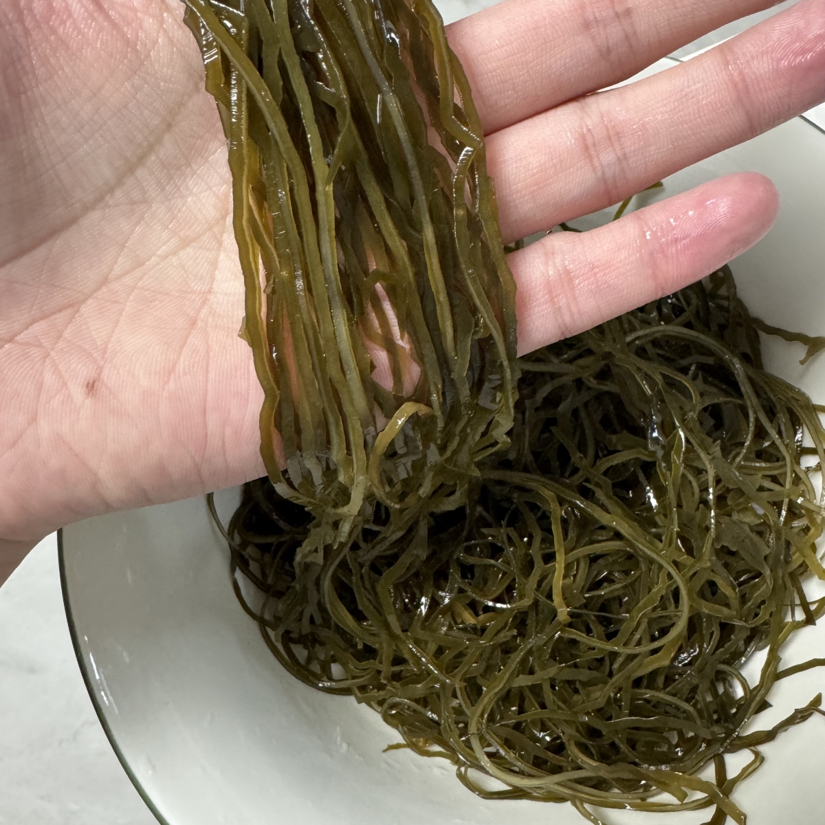 一位女性在现代厨房中操作,戴着围裙,右手持不锈钢漏勺从冒着热气的锅中捞出焯好水的深褐色海带丝,锅边放有生姜片和料酒瓶,背景清晰体现家庭烹饪环境,突出安全卫生的操作过程 厨房操作场景:女性手部正在用漏勺捞出沸水中的海带丝