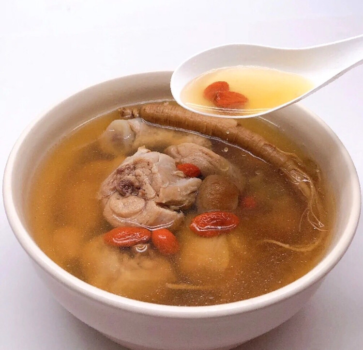 一道精致的陶瓷碗盛装的生晒参炖鸡汤,汤色清澈微黄,可见两片完整的生晒参横卧其中,周围点缀几颗枸杞和葱花,旁边摆放一双筷子和木制餐垫,体现传统中式养生饮食美学 生晒参烹饪菜肴成品图