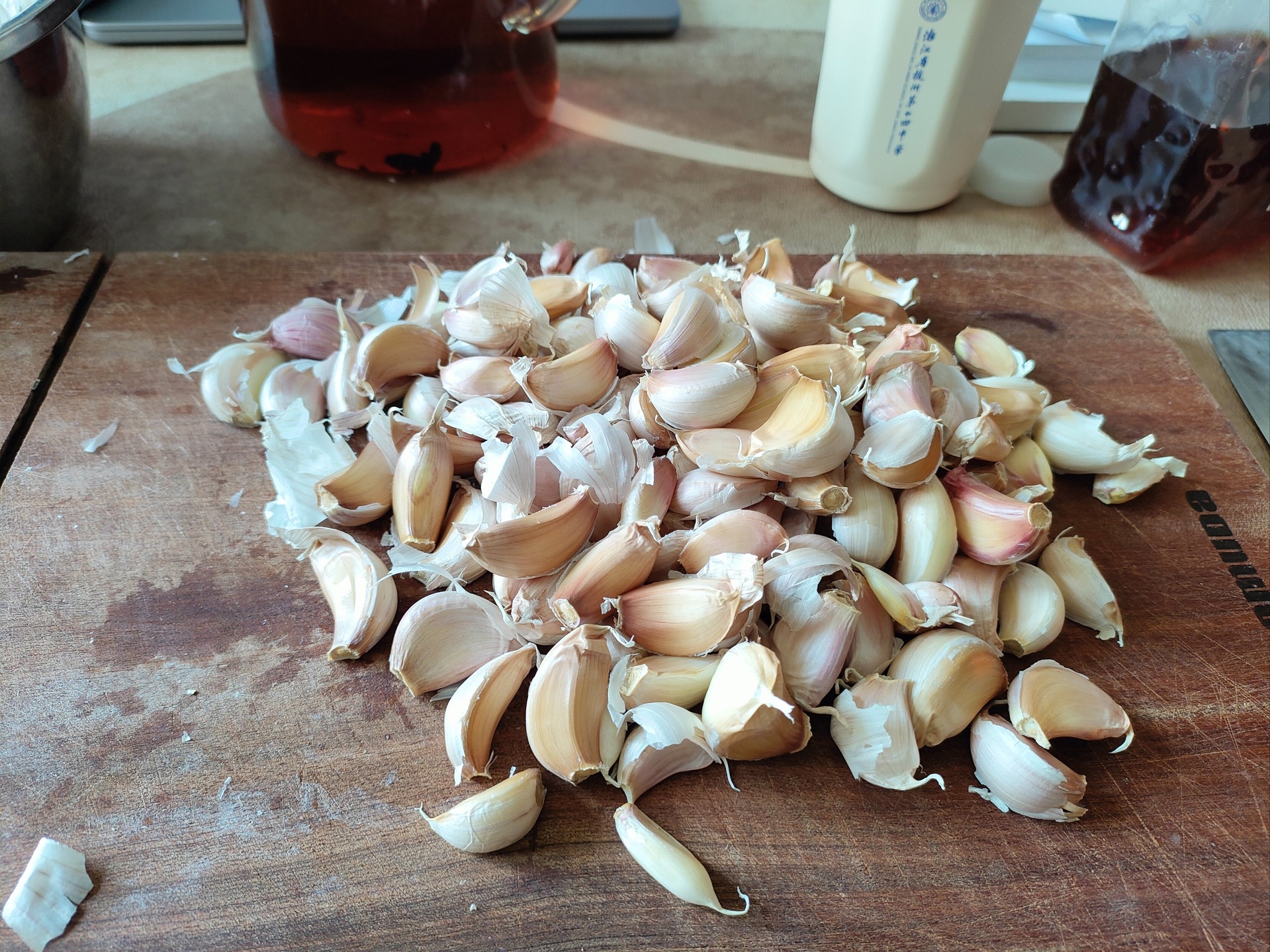 一名穿着围裙的厨师在光线明亮的厨房内使用木制擀面杖在石臼中捣碎新鲜大蒜,石臼内呈现湿润的乳白色蒜泥,背景桌上摆放着几根已蒸熟去蒂的紫皮长茄子,准备用于制作凉拌菜 一位厨师正在厨房操作台上用擀面杖捣碎大蒜瓣,石臼中可见白色蒜泥,旁边放着蒸好的茄子