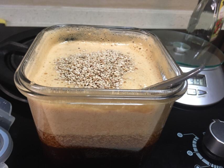 一位厨师正在玻璃杯中用打蛋器搅拌球芝麻预粉拌,杯子中有半稠状液体,手部动作显示顺时针搅拌,桌面上放有量勺、蜂蜜瓶和温水壶,展示完整冲调流程 芝麻糊冲调过程