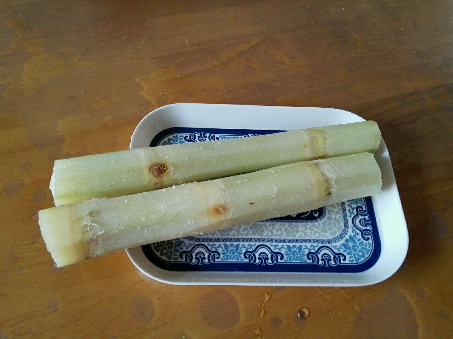 一位女性在厨房操作台上用刀将一根细黑皮甘蔗切成小段，旁边放着榨汁机和玻璃杯，桌上铺有木质砧板和绿色抹布