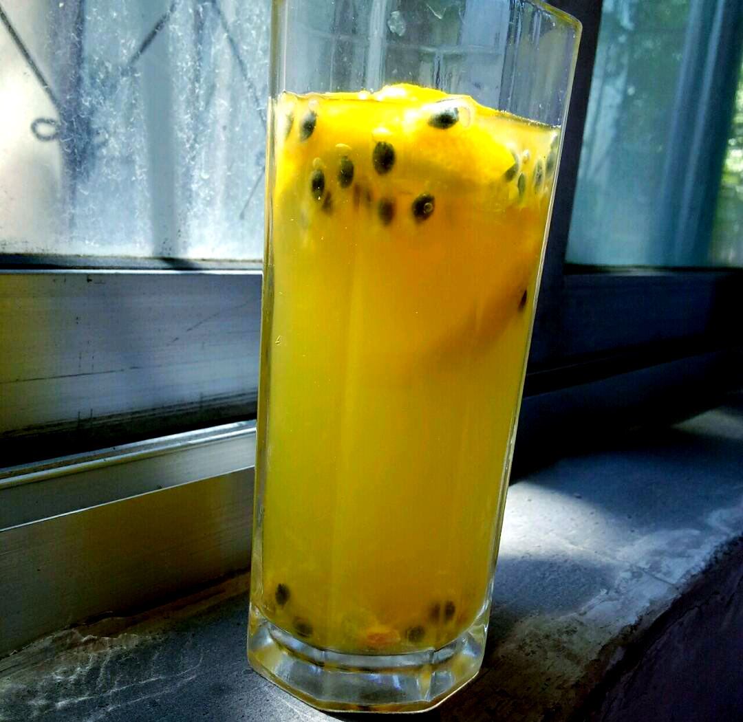 一杯透明玻璃杯中盛有淡黄色果汁饮料,内含可见的黄金百香果籽粒,杯壁点缀着半个切开的黄金百香果,旁边摆放一碟切好的新鲜水果,整体风格清新自然,适合夏季饮用场景 黄金百香果饮品搭配图