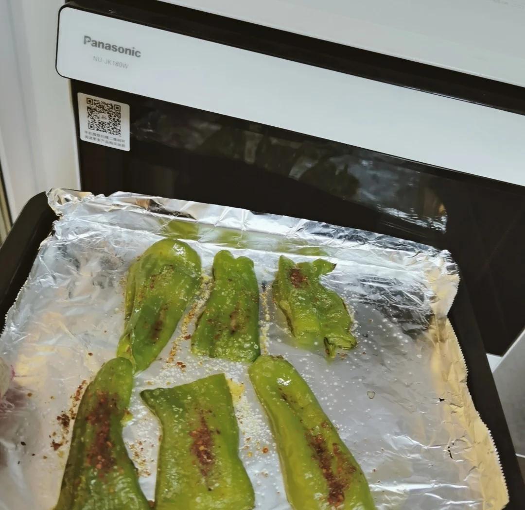 一个不锈钢烤盘上摆放着三只正在烘烤的青圆椒,表面已经出现明显的焦斑和鼓泡现象,背景为开启状态的家用烤箱,温度显示220摄氏度,旁边有一把硅胶刷沾着金色橄榄油 烤青圆椒制作过程展示