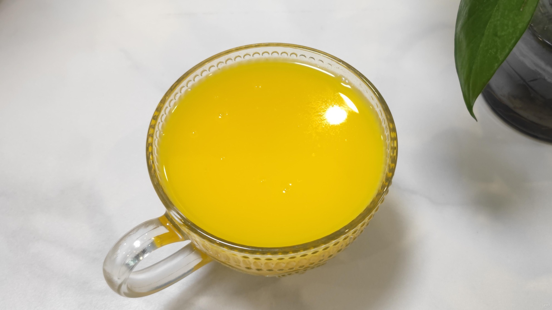 一位手部特写正端着一杯透明玻璃杯装的金桔柠檬饮品,杯中有清晰可见的金桔块和柠檬片,冰块晶莹,液体呈淡黄色,背景为浅色木桌与绿植 手握透明饮品杯展示金桔柠檬茶