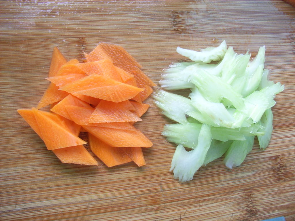 新鲜胡萝卜与芹菜摆放在木质砧板上，旁边有榨汁机和玻璃杯，背景为自然光线厨房环境