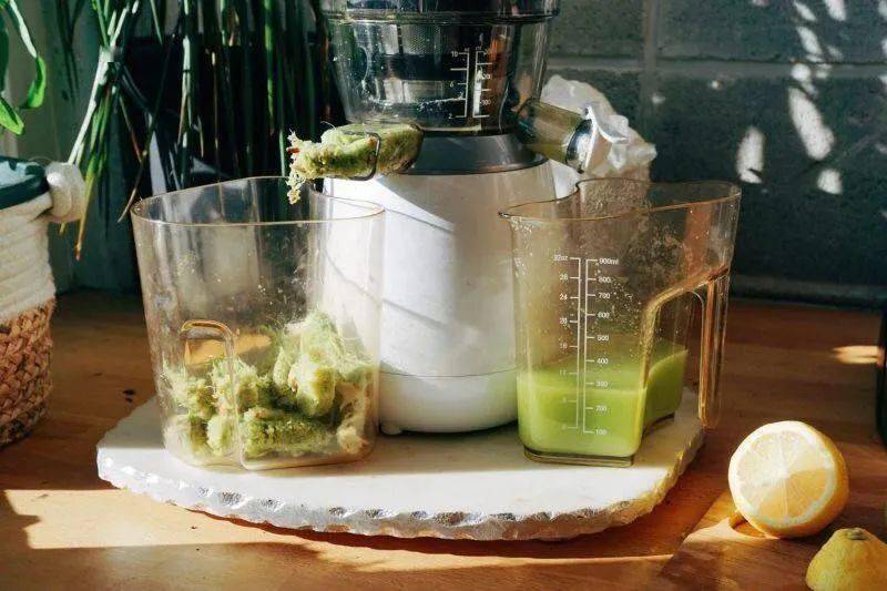 一位女性在明亮现代的厨房操作台上正在用玻璃杯调制柠檬饮品,手握榨汁器挤压柠檬片,旁边放有蜂蜜罐、量勺、冰块托盘和切好的柠檬片,桌面整洁温馨,突出健康生活方式氛围 家庭厨房中使用安岳柠檬制作饮品的场景