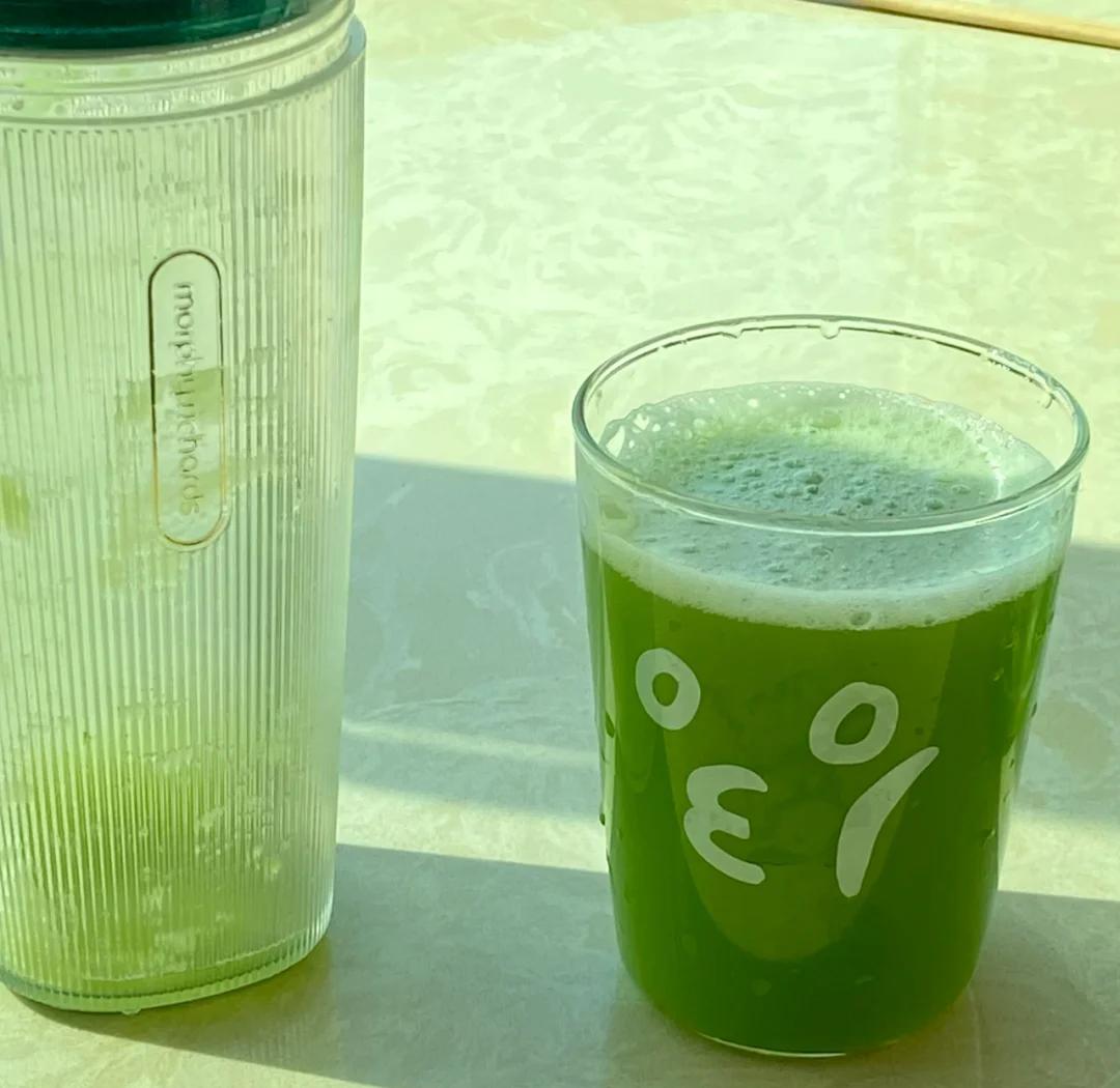 一位女性坐在阳光充足的餐桌前,手持绿色果汁微笑饮用,桌上放有菠菜、水果和食谱笔记,营造健康生活方式氛围 图片描述