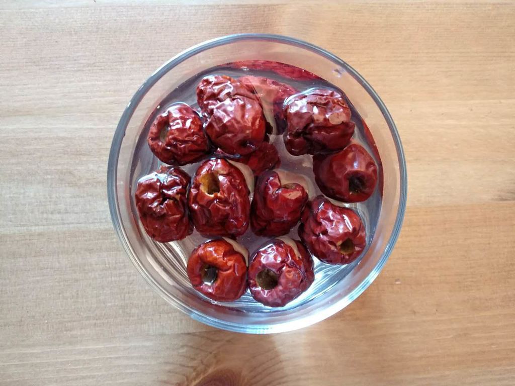 一名厨师正在厨房操作台上将干桂圆和干红枣片倒入透明玻璃碗中,旁边有流动的清水,手部动作显示正在进行轻柔冲洗。背景整洁明亮,体现食品安全与卫生细节 干桂圆与干红枣片清洗浸泡过程