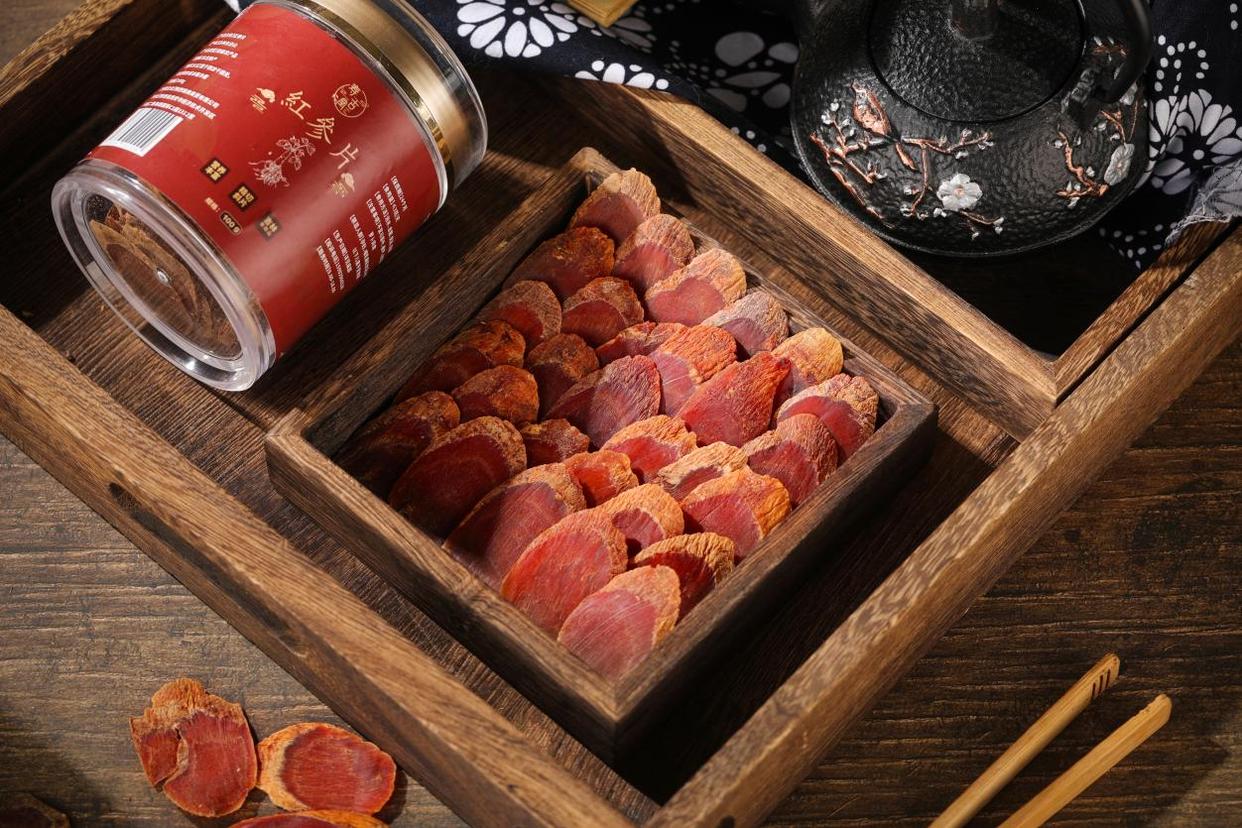 画面左侧为干燥的红参切片,呈深红褐色,表面有明显纹理;右侧为透明玻璃杯中正在用热水冲泡的红参片,水中泛起淡淡的金黄色,部分红参片漂浮,背景为木质茶桌和传统中式茶具,营造出养生饮茶氛围 红参切片与热水冲泡过程对比图