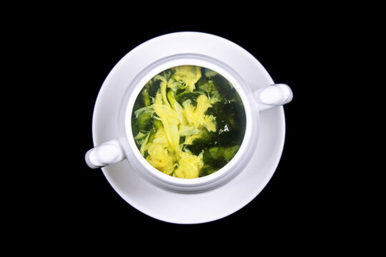 白色瓷碗中盛放着碧绿清澈的绿豆海带鸡蛋汤,蛋花如云朵般漂浮,点缀着翠绿葱花,旁边摆放木勺和竹筷,背景为简约餐桌布景 成品展示图