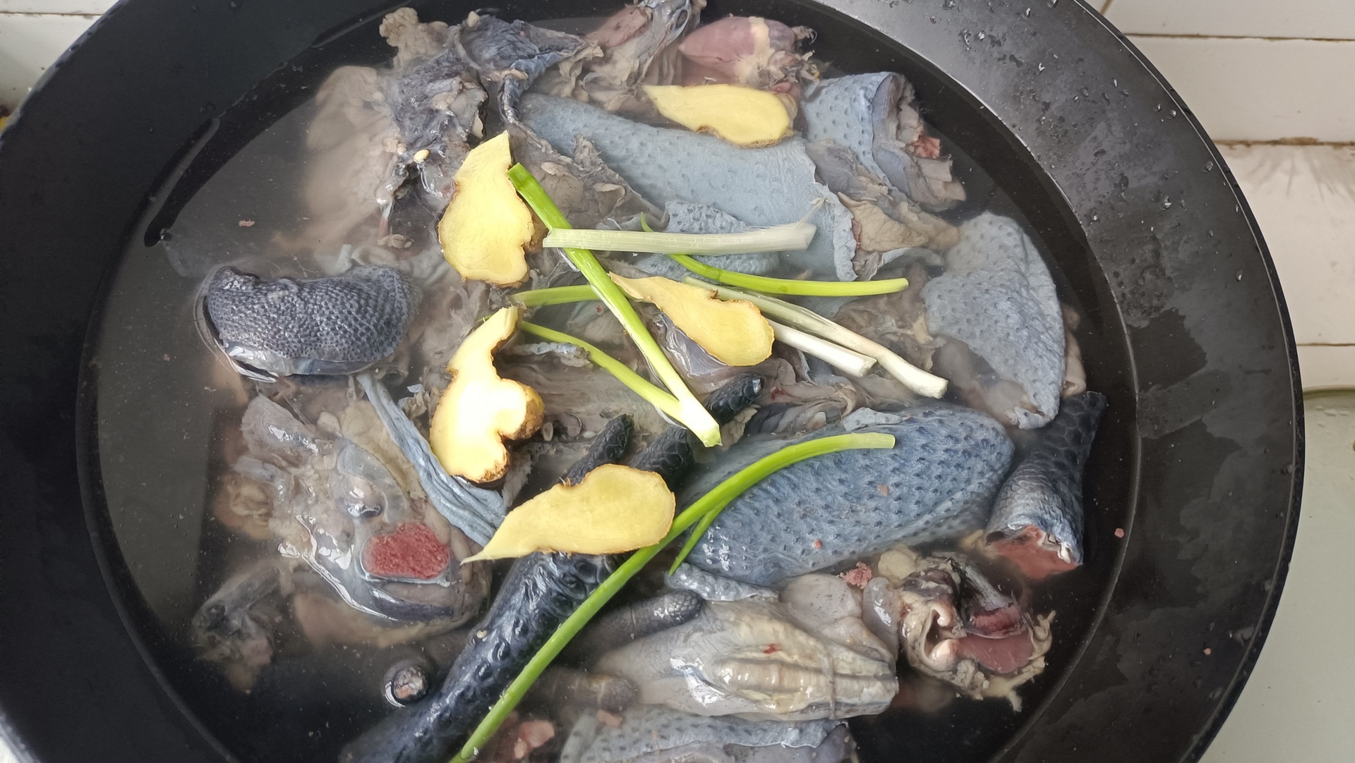 一只乌鸡块放入冷水锅中,水面刚没过鸡肉,旁边摆放着几片生姜和一瓶料酒,炉火微燃,水尚未沸腾,用于展示焯水初始阶段 乌鸡焯水过程特写