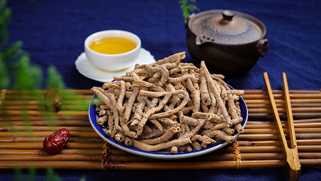 一名身穿传统中式服装的中医师在古色古香的诊室内操作,桌上摆放着木质药柜、铜秤、瓷罐,手中拿着研钵正在研磨药材,旁边放着十颗完整的长枣,背景墙上挂着‘十枣汤’字样的书法作品 一位中医师正在研磨药材并放入陶罐中煎煮