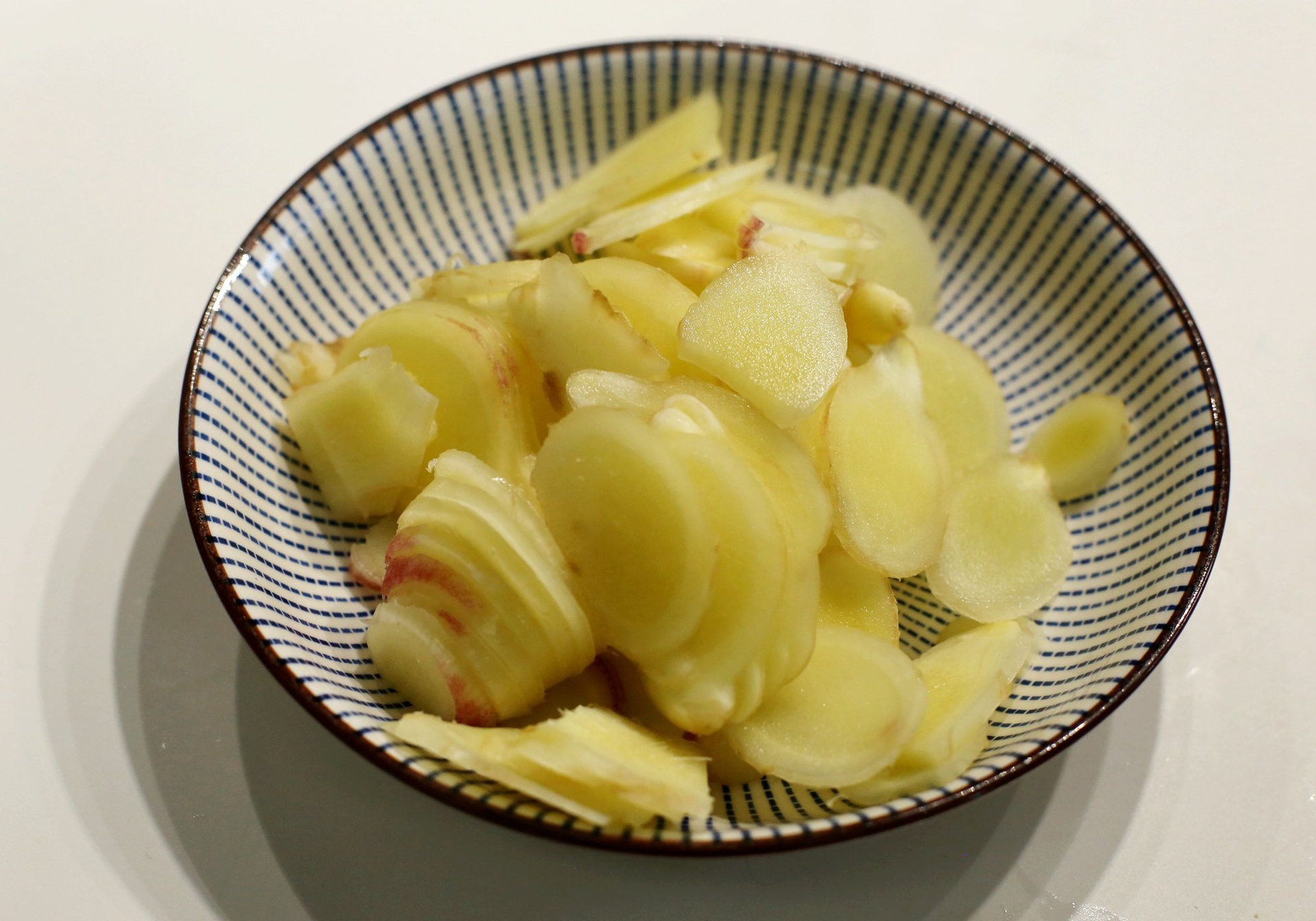 一位中年妇女在厨房灶台前熬制姜汤，左手拿着姜块正在切片，右手边是装有生姜的竹篮，灶上的铁锅冒着热气，墙上挂着老式铜勺和陶罐