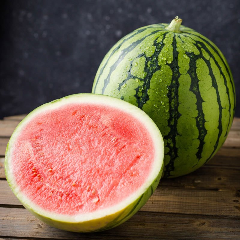 一个完整的西瓜放在木桌上,旁边是切开后的两半西瓜,其中一半呈现鲜艳的红色果肉,另一半则显示淡黄色或浅粉色的果肉,形成鲜明对比,背景为自然光下的厨房环境 西瓜切开对比图