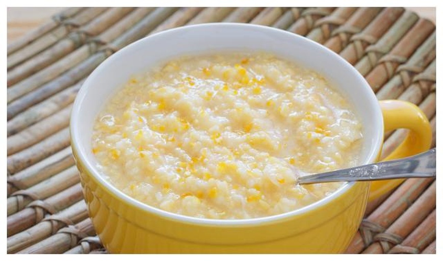 一碗热气腾腾的雪梨小米粥，粥体呈淡黄色，表面可见细小的雪梨丁，质地细腻顺滑，盛放在白色瓷碗中，背景为木质餐桌和秋季落叶装饰