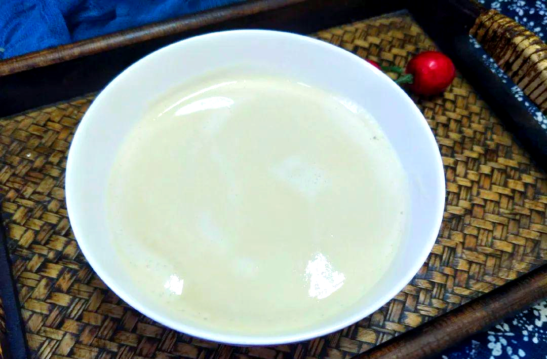 一只白色陶瓷杯中盛满乳黄色的花生豆浆,表面泛着细腻光泽,靠近可见微小气泡均匀分布。杯子置于木质餐垫上,旁边放有一把不锈钢勺子和一小碟红糖,背景虚化,营造温馨早餐氛围 成品花生豆浆特写