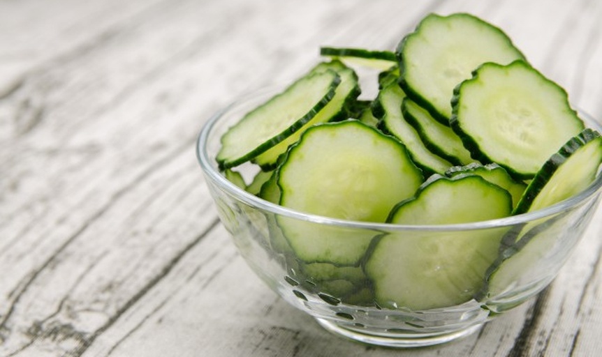 一杯透明玻璃杯中盛满浅绿色的新鲜果蔬汁,表面有细微气泡,背景为切开的黄瓜片和新鲜生菜叶散落在木质料理台上,自然光线照射,体现清新健康的视觉氛围 清新果蔬汁特写