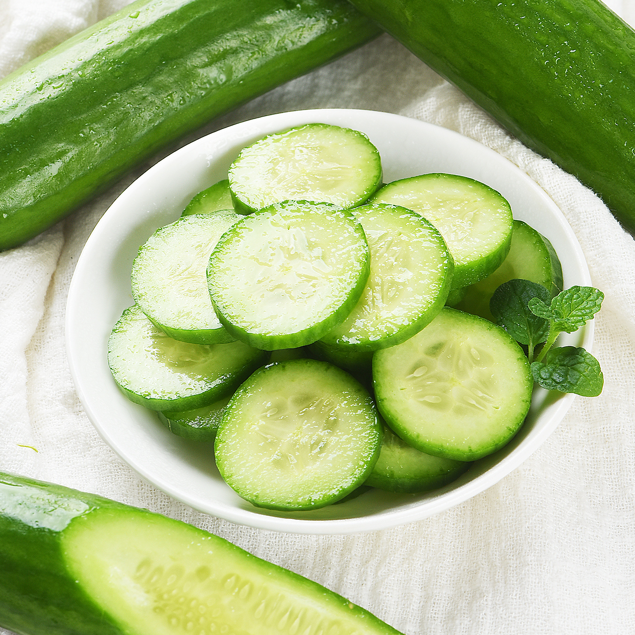 一名穿着围裙的女性正在处理新鲜黄瓜小扭,将其切成圆形薄片,案板上散落着翠绿瓜片和黄色柠檬片,背景有透明玻璃容器和不锈钢厨具,营造现代健康饮食氛围 一位女性厨师正在厨房操作台上将新鲜小黄瓜切成薄片,旁边配有柠檬片、橄榄油瓶和玻璃沙拉碗,整体色调清新明亮