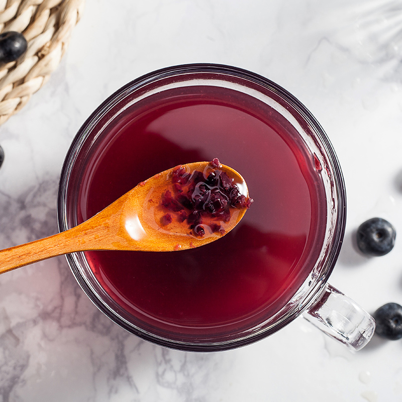 一名女性手持玻璃杯品尝自制蓝莓果蔬汁，面带微笑，背景为阳光照射的厨房台面，体现健康生活方式