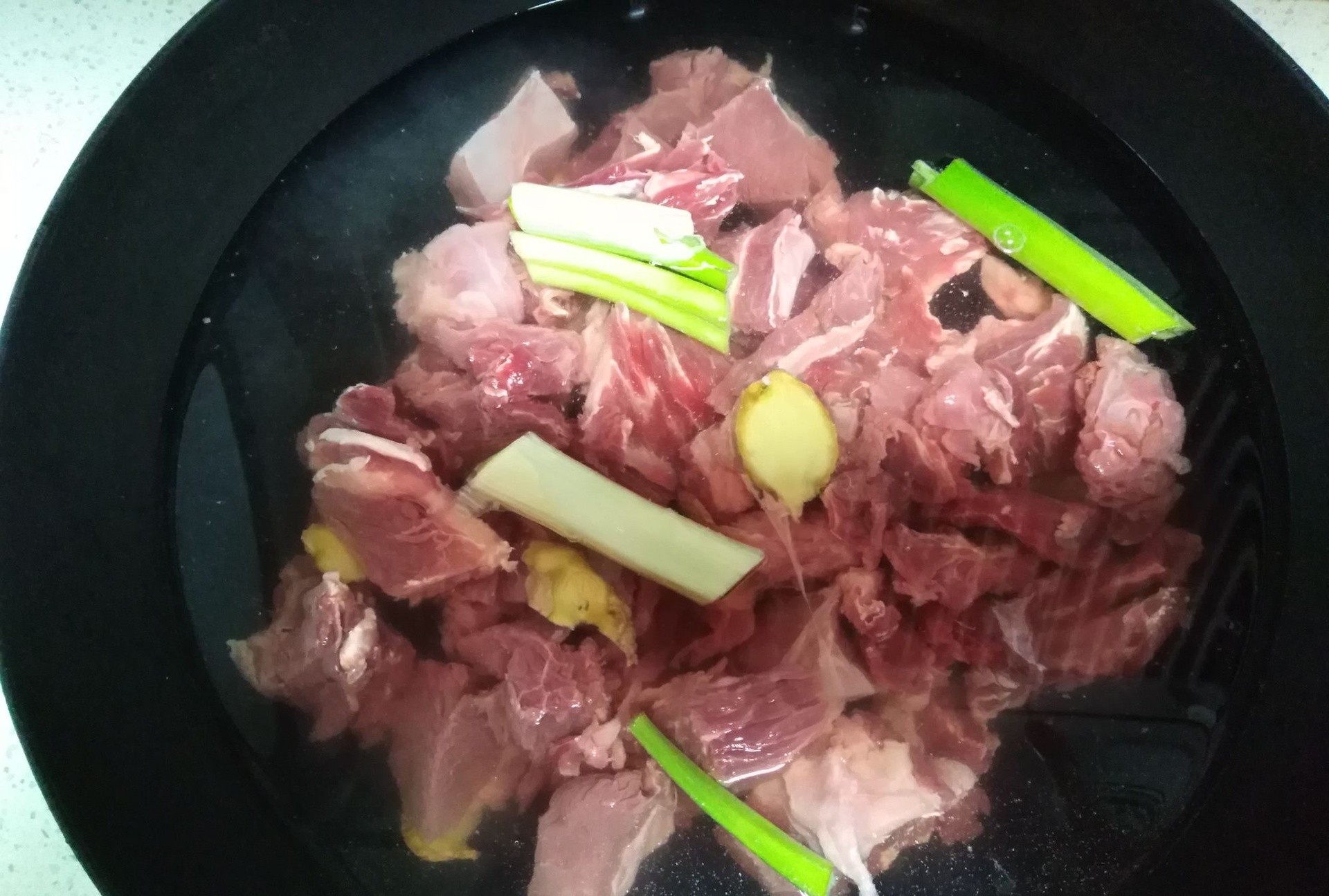 一块块切好的瘦肉块放入冷水中,锅边已出现灰白色浮沫,旁边摆放着姜片和一瓶料酒,正在用勺子轻轻撇去浮沫 瘦肉焯水过程