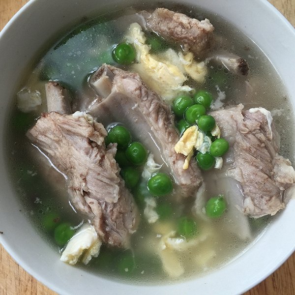 一碗热气腾腾的豌豆排骨汤,汤色清亮微白,碗中有大颗绿色豌豆、小块排骨和胡萝卜丁,旁边摆放一双筷子和一个陶瓷勺,突出家庭养生饮食氛围 豌豆排骨汤成品图