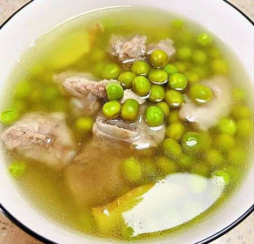 一位中老年女性正在厨房灶台前用砂锅炖煮豌豆排骨汤,锅中汤色乳白,飘有绿色豌豆和胡萝卜块,体现豌豆作为药膳食材的家庭应用场景 老年人在厨房中煮豌豆汤