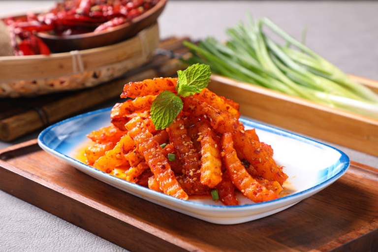 一个不锈钢大碗中盛放着刚炸好的金黄色狼牙土豆条,厨师正用夹子将热油泼向碗中的辣椒面和蒜泥,瞬间腾起香气,调料开始融合,土豆表面已部分裹上红亮油汁 狼牙土豆调味拌制过程