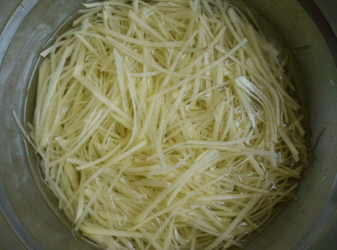 切好的土豆丝浸泡在透明玻璃碗的清水中，水面略高于土豆丝，旁边放着一把厨房剪刀和半个柠檬，背景为浅色厨房台面