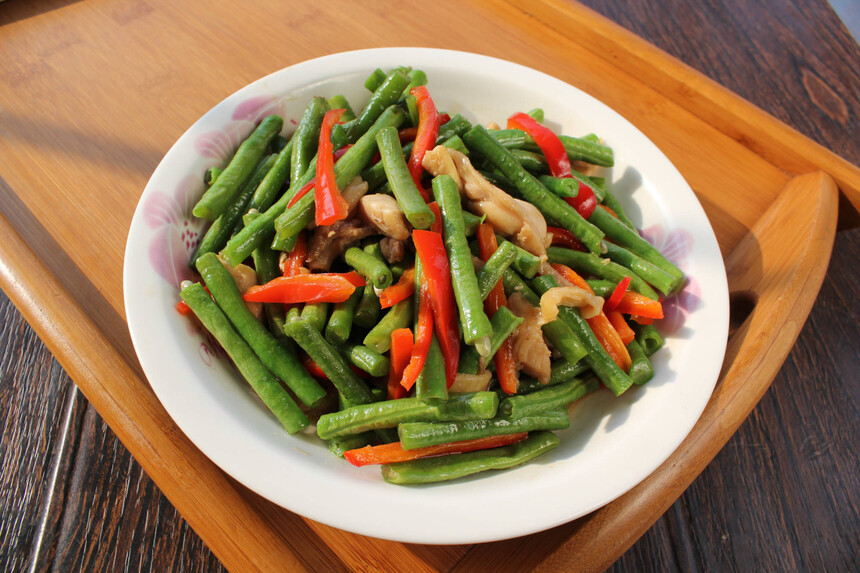 泡椒炒四季豆烹饪过程关键帧：左手持锅铲翻动油中四季豆与泡椒，右手持小碗正沿锅边淋入琥珀色调味汁，锅内红绿交织、热气升腾，灶火呈蓝色稳定火焰