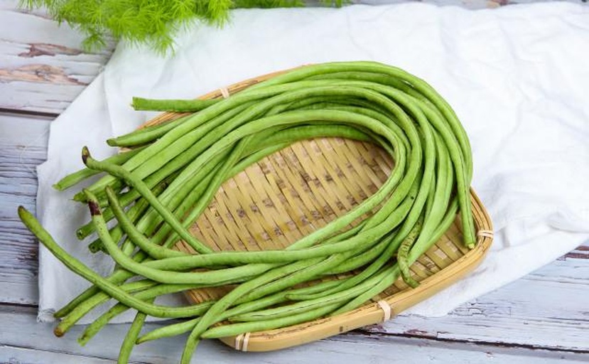 两个竹编篮子分别盛放新鲜豇豆与菜豆:左侧篮中为数十根修长翠绿的豇豆,整齐捆扎;右侧为短粗饱满的浅绿色菜豆,部分带有淡紫色花斑。背景为清晨农贸市场场景,光线柔和,体现新鲜度与地域特色 新鲜采摘的豇豆与菜豆分筐陈列图