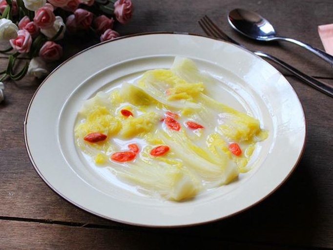 一盘热气腾腾的上汤娃娃菜,金黄色的汤底中浸泡着洁白嫩绿的娃娃菜段,表面点缀有红色枸杞、黄色胡萝卜片和少许蒜蓉,菜品色泽鲜艳,汤汁清澈透亮,摆盘精致,背景为浅色陶瓷餐盘与木质餐桌 上汤娃娃菜成品特写