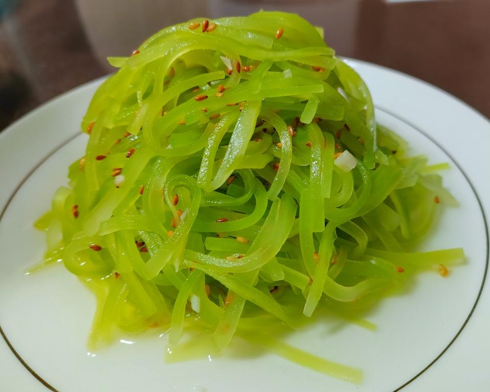 俯拍视角:晶莹莴苣丝堆叠成小山状,表面均匀裹覆琥珀色酱汁,可见细密蒜末、红亮小米辣圈与黑白芝麻点缀,背景为青花瓷浅盘,丝体根根分明无黏连 凉拌莴苣丝全景