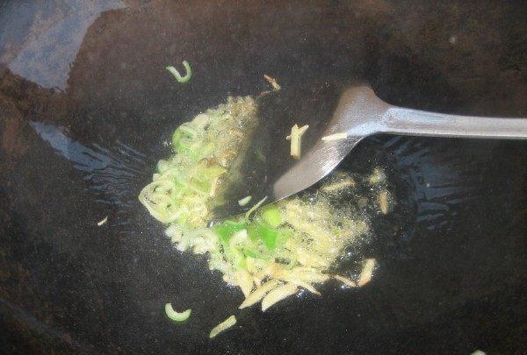 锅中正在煸炒姜蒜葱段，油面泛起金黄色光泽