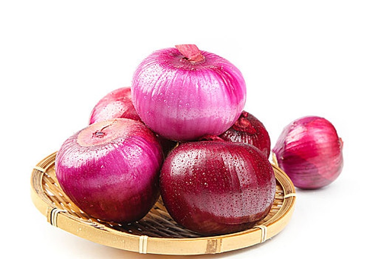 红皮洋葱特写：整颗带根须的新鲜红皮洋葱，表皮呈均匀紫红色，略带蜡质光泽，部分外层鳞片微翘，切面可见粉白相间的同心环状结构，背景为木质砧板与阳光洒落的自然光效