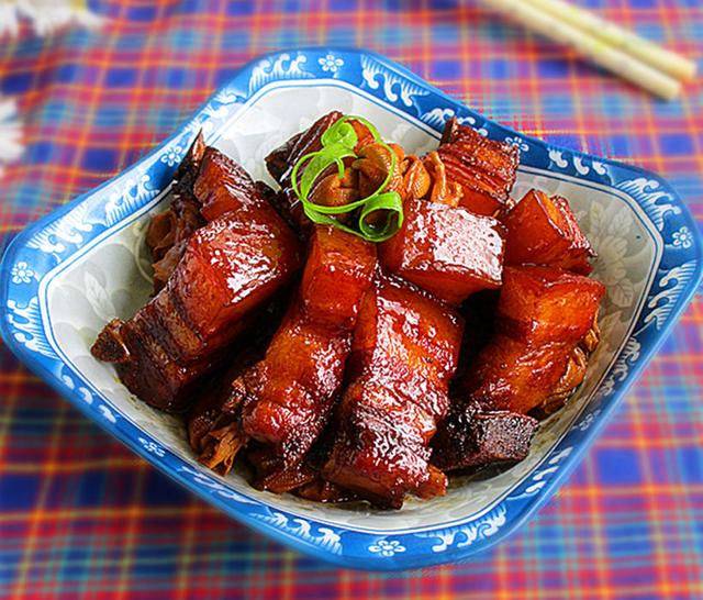 一盘色泽红亮的红烧肉摆放在白色瓷盘中,表面油润反光,旁边点缀绿色葱花,背景虚化的灶台上可见使用的八角和调料瓶 成品红烧肉装盘效果