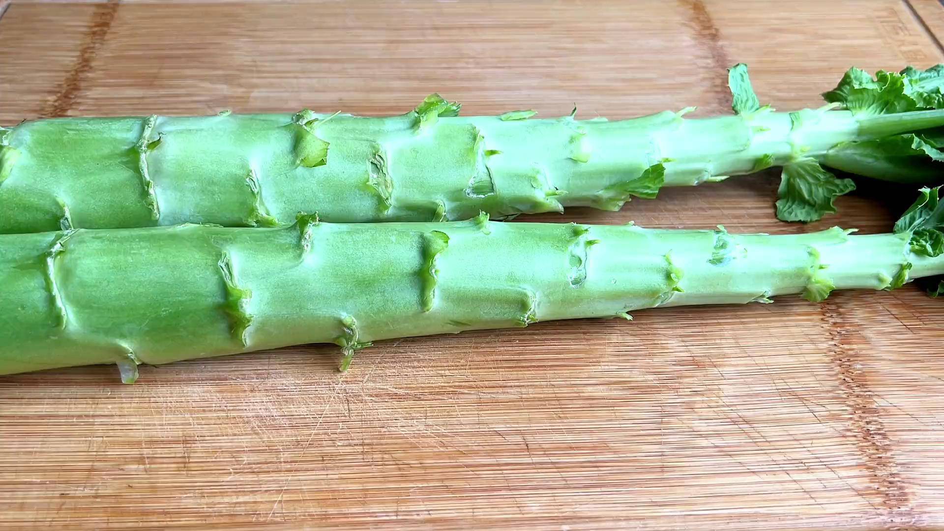 一根表皮鲜绿、带有细密纵纹的新鲜莴苣平放在木质案板上,顶部保留少量叶片,刀具在一旁准备削皮,背景为厨房自然光环境 新鲜莴苣的外观特征