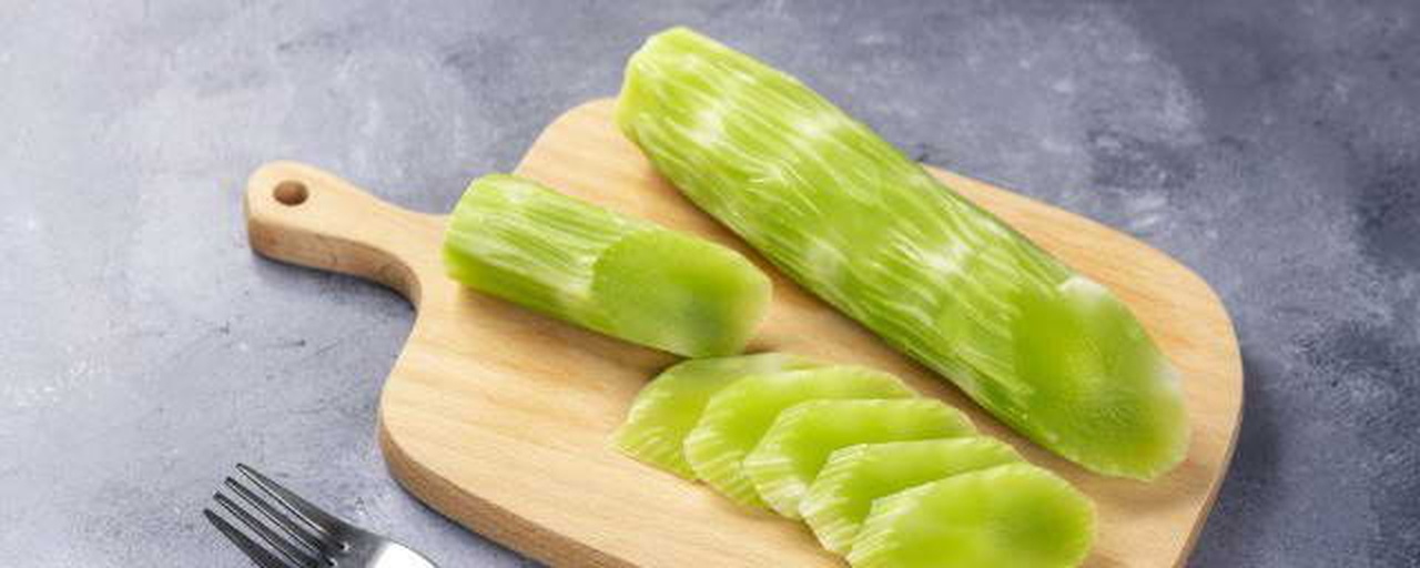 一个明亮的家庭厨房操作台上,摆放着已削皮切片的莴苣笋,旁边有切好的红椒丝、蒜末和一个小碗中的酱油调料,锅中正在翻炒食材,散发出轻烟,背景可见木质橱柜和绿植 家庭厨房中的莴苣笋料理场景