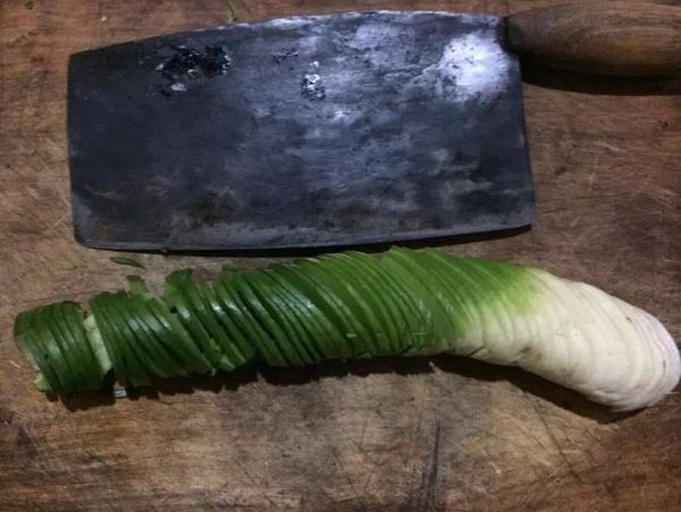 一名厨师正在案板上对一根新鲜的章丘大葱进行斜刀切割,旁边有清水冲洗过的葱段和剥下的外皮,背景为厨房操作台,光线明亮,突出食材的新鲜与洁净 章丘大葱的清洗与切割过程