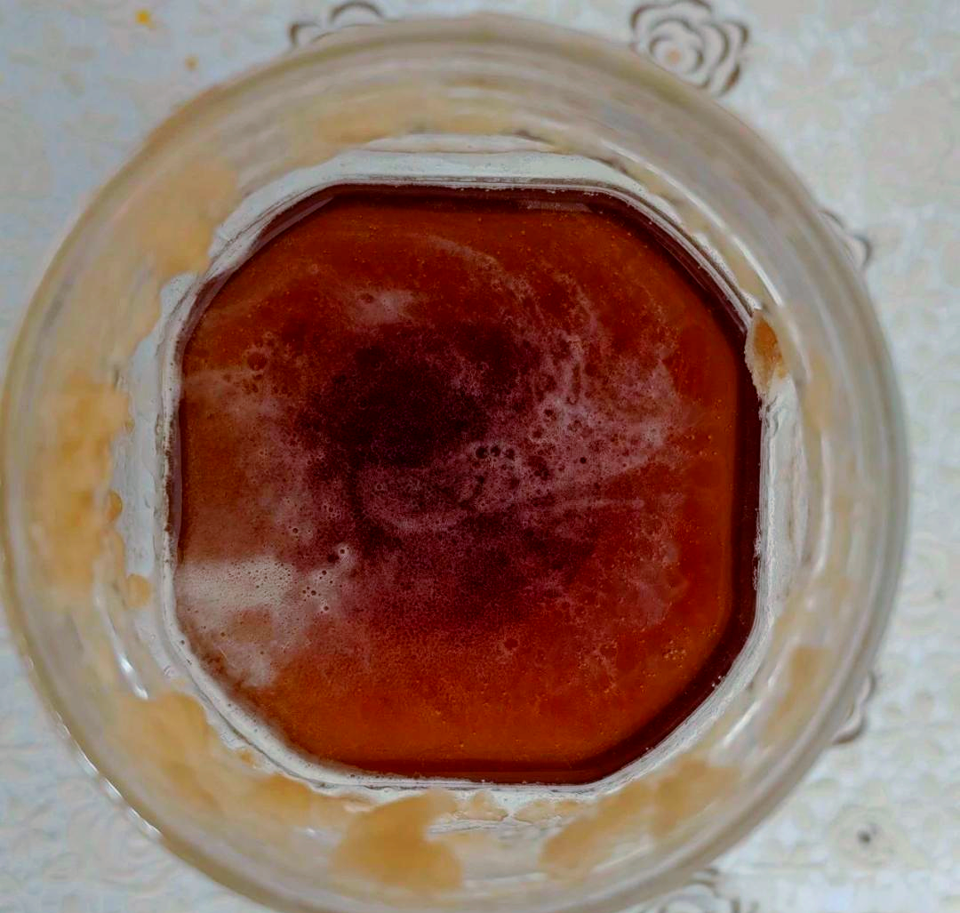 一个透明玻璃发酵罐内装有淡黄色液体,可见细密气泡缓缓上升,罐口连接着水封装置,水面有小气泡冒出,周围环境整洁,背景为木质架子 玻璃发酵罐中的黄桃果酒发酵过程