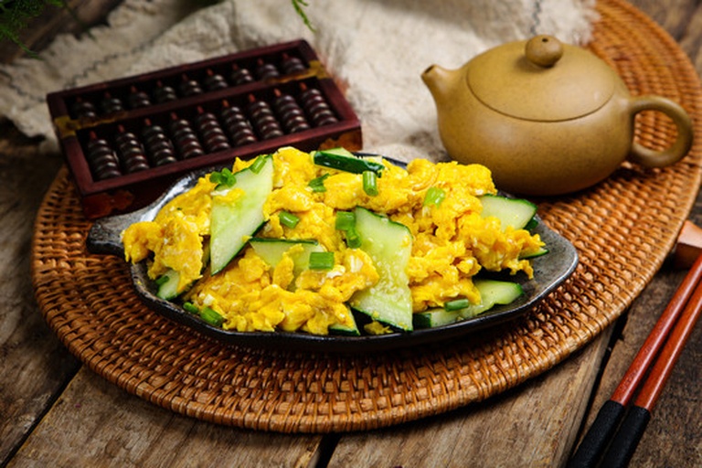 成品小黄瓜炒鸡蛋盛放在白色瓷盘中，色泽金黄与翠绿相间，旁边配有米饭和竹筷